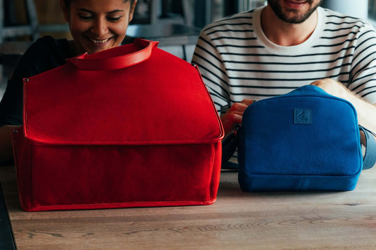 Deux cabas en velours, un rouge et un bleu, présentés sur une table avec des personnes souriantes.